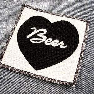 May include: A white fabric patch with black stitching around the edges. A black heart with the word "Beer" written in white inside the heart is centered on the patch.