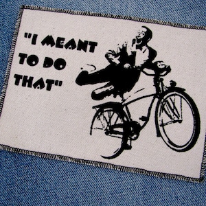 May include: A white patch with black text and a black and white illustration of a person riding a bicycle. The text reads "I meant to do that".