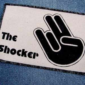 May include: A white patch with black stitching around the edges. The patch has a black and white image of a hand making the "shocker" gesture. The text "The Shocker" is printed in black below the image.