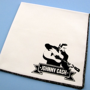 May include: White cotton handkerchief with black embroidered image of Johnny Cash playing a guitar. The text "JOHNNY CASH" is on a banner below the image.