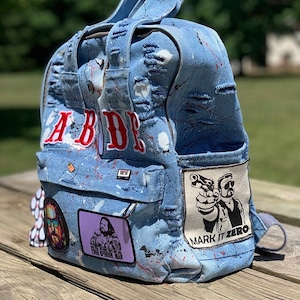 May include: A blue denim backpack with white paint splatters and patches. The backpack has a large white patch with the text "Mark It Zero" and a black and white image of two men holding guns. The backpack also has a smaller patch with the text "Your Opinion, Man" and a black and white image of a man. The backpack has the word "ABIDE" in red letters on the front.