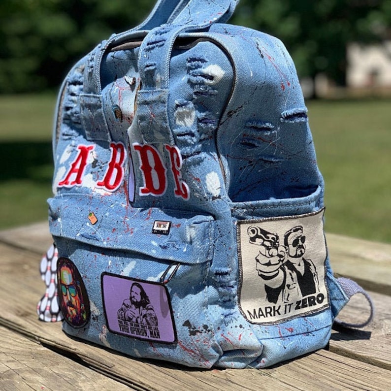 May include: A blue denim backpack with white paint splatters and patches. The backpack has a large white patch with the text "Mark It Zero" and a black and white image of two men holding guns. The backpack also has a smaller patch with the text "Your Opinion, Man" and a black and white image of a man. The backpack has the word "ABIDE" in red letters on the front.