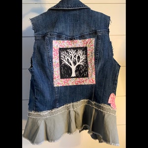 May include: A blue denim vest with a floral fabric patch featuring a black tree design. The vest has a ruffled bottom and a pink heart-shaped patch.