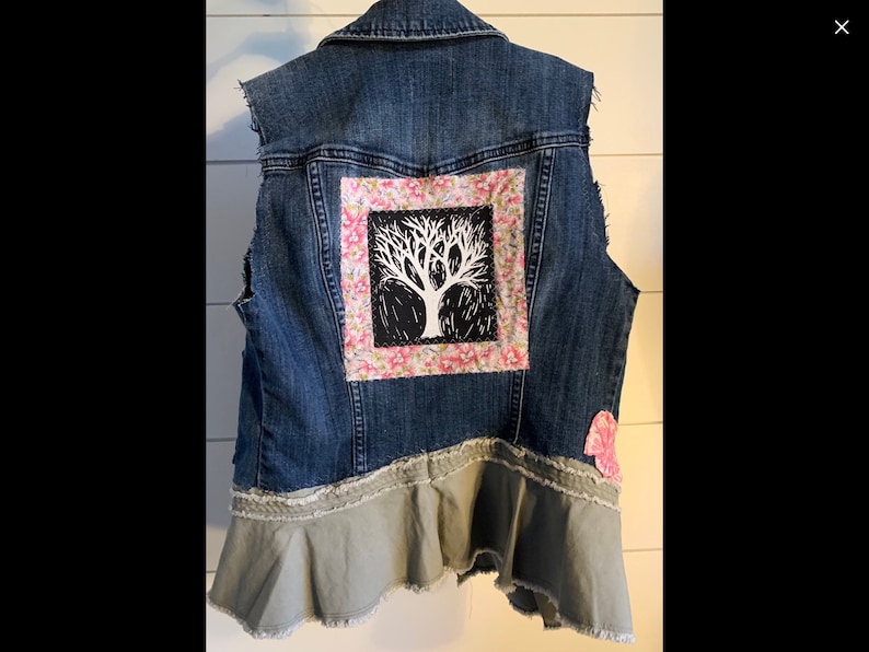 May include: A blue denim vest with a floral fabric patch featuring a black tree design. The vest has a ruffled bottom and a pink heart-shaped patch.