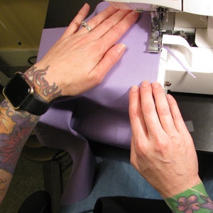 May include: A person's hands are shown sewing a piece of purple fabric on a sewing machine. The person is wearing a watch and has tattoos on their arm.