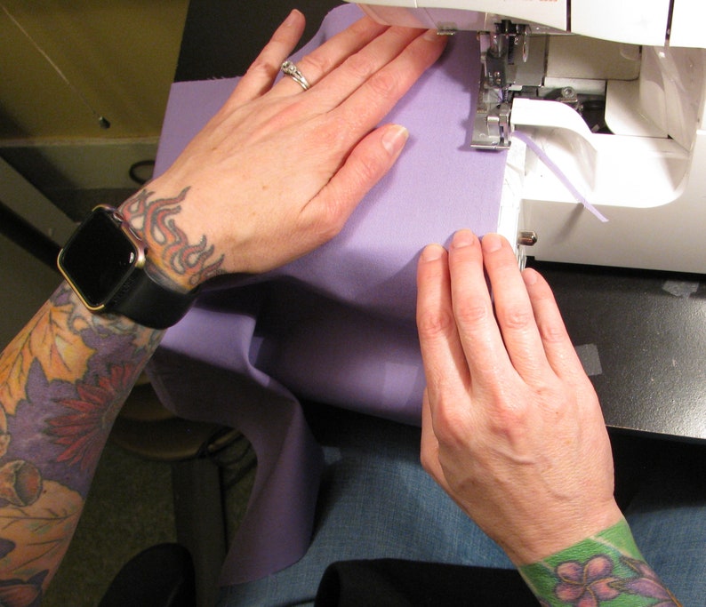 May include: A person's hands are shown sewing a piece of purple fabric on a sewing machine. The person is wearing a watch and has tattoos on their arm.