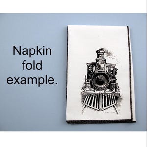 May include: A white napkin with a black and white illustration of a vintage steam locomotive. The napkin is folded in half and has a black border.