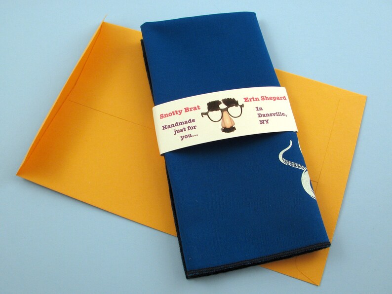 May include: A blue fabric napkin with a white octopus design is wrapped with a white label that reads "Snotty Brat Handmade just for you... Erin Shepard In Dansville, NY".