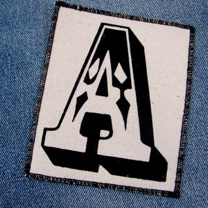May include: A black and white fabric patch with a stylized letter 'A' in a bold, graphic font.