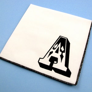May include: White fabric square with a black, stylized letter A printed on it. The fabric has a black stitched border.