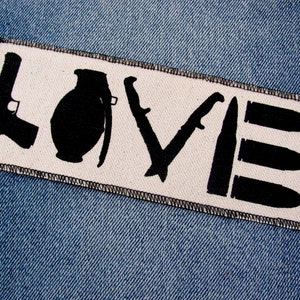 May include: A rectangular patch with the word "LOVE" spelled out using weapon silhouettes. The letters are formed by a gun, grenade, knife, and bullets. The patch is white with black stitching and is set against a blue denim background.
