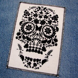 May include: A black and white floral skull design printed on a white fabric patch. The patch is sewn onto a blue denim background.
