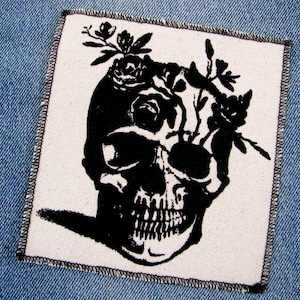 May include: Black and white embroidered patch of a skull with flowers growing out of the top of its head.