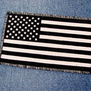 May include: Black and white American flag patch with a stitched border. The patch is on a blue denim background.