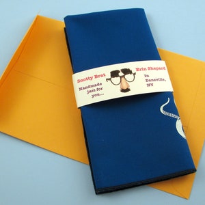 May include: A blue fabric napkin with a white octopus design is wrapped with a white label that reads "Snotty Brat Handmade just for you... Erin Shepard In Dansville, NY".