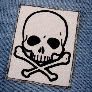 May include: A white fabric patch with a black and white skull and crossbones design. The skull has two black eye sockets and a black mouth.