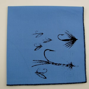 May include: A light blue fabric napkin with a black print of various fishing flies. The flies are depicted with detailed hooks and feathers.