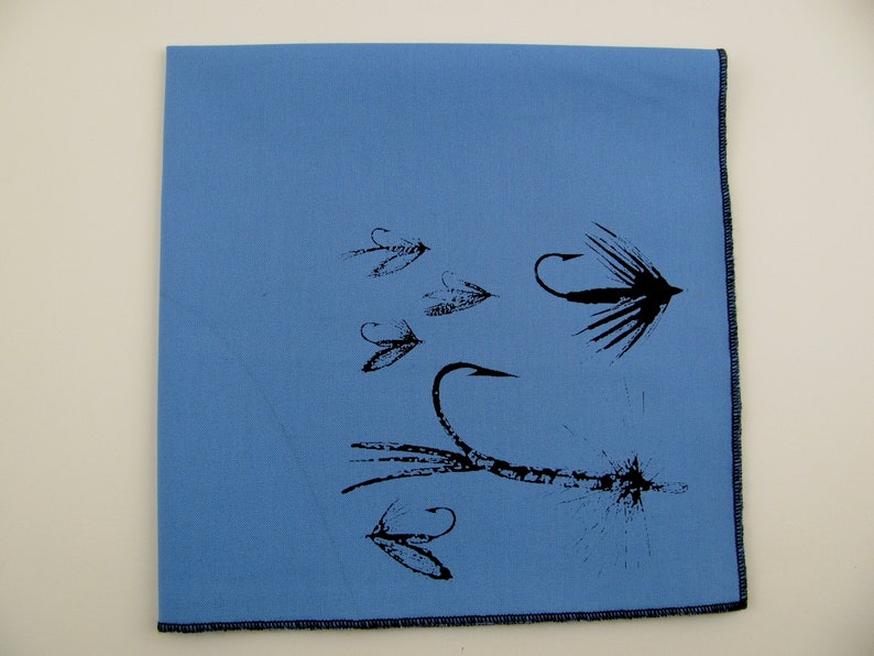 May include: A light blue fabric napkin with a black print of various fishing flies. The flies are depicted with detailed hooks and feathers.