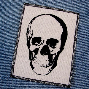 May include: Black and white skull patch with a stitched border. The skull is printed on a white fabric background.