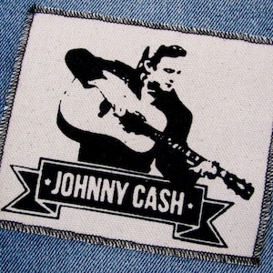 May include: Black and white patch with a silhouette of Johnny Cash playing a guitar. The patch has a banner with the text "JOHNNY CASH" in black.