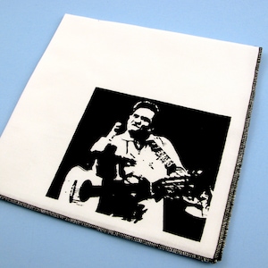 May include: Black and white image of a man playing a guitar. The man is wearing a button-down shirt and has a microphone in his hand. The image is printed on a white fabric.
