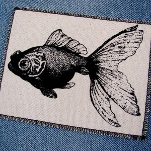 May include: Black and white illustration of a goldfish with a large, round eye. The fish is printed on a white fabric patch with a black stitched border.