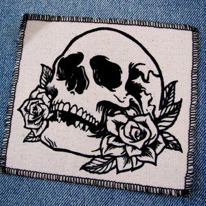 May include: Black and white embroidered patch of a skull with a rose in its mouth. The skull is outlined in black and the rose is detailed with petals and leaves.