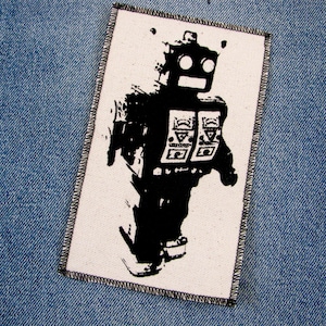 May include: Black and white iron-on patch featuring a robot with a simple design. The robot has two large eyes and a rectangular body with a few details.