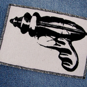 May include: A rectangular patch featuring a black ray gun design on a white canvas. The patch is edged with black stitching and is displayed on a blue denim background.