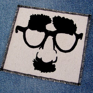 May include: Black and white illustration of a person wearing glasses and a mustache. The illustration is on a white fabric background.