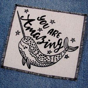 May include: A white fabric patch with black stitching around the edges. The patch features a black and white illustration of a narwhal with a unicorn horn and the text "You are amazing" with stars.