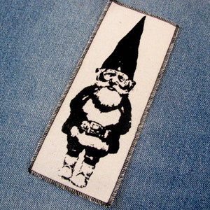 May include: Black and white iron-on patch of a cartoon gnome wearing a pointed hat and a belt. The gnome is standing with its arms crossed and has a serious expression.