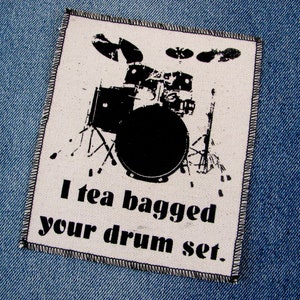 May include: A white fabric patch with black text and a black silhouette of a drum set. The text reads "I tea bagged your drum set."