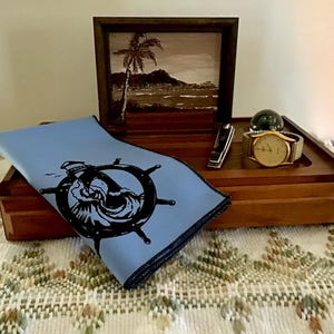 May include: A wooden box with a blue cloth inside. The cloth has a black and white image of a ship's wheel with waves. A gold watch and a silver pocket knife are on top of the box.