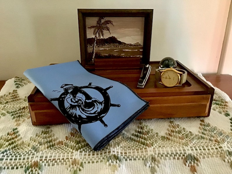 May include: A wooden box with a blue cloth inside. The cloth has a black and white image of a ship's wheel with waves. A gold watch and a silver pocket knife are on top of the box.