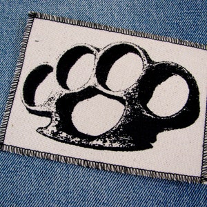 May include: Black and white iron-on patch of a brass knuckle. The patch is on a blue denim background.