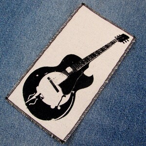 May include: A rectangular patch featuring a black and white illustration of an archtop guitar. The guitar is depicted in a silhouette style against a white background. The patch is set against a blue denim background.