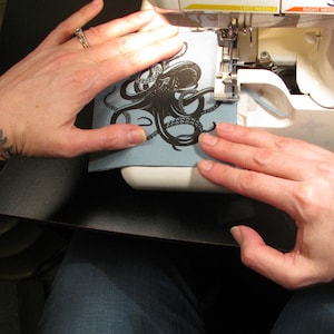 May include: A person's hands are holding a blue fabric patch with a black octopus design in front of a white sewing machine.