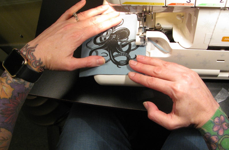 May include: A person's hands are holding a blue fabric patch with a black octopus design in front of a white sewing machine.