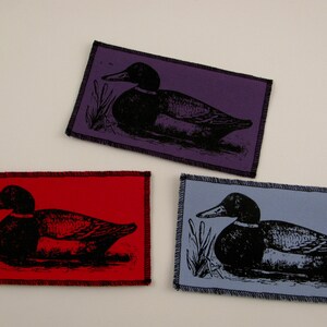 May include: Three fabric patches with black and white images of ducks on different colored backgrounds. The patches are purple, red, and blue.
