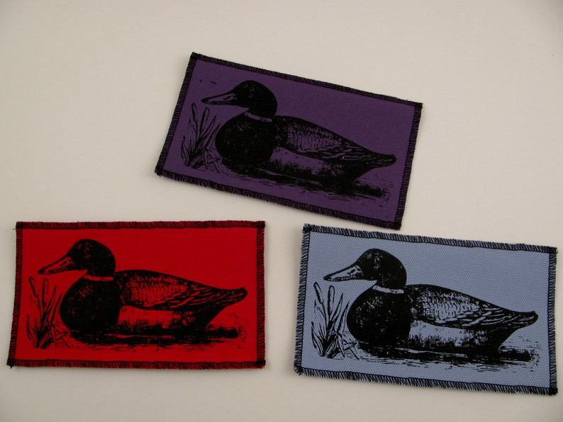 May include: Three fabric patches with black and white images of ducks on different colored backgrounds. The patches are purple, red, and blue.
