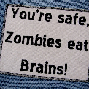 May include: A white patch with black text that reads "You're safe, Zombies eat Brains!" The patch is sewn onto a blue denim background.