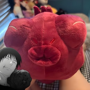 Peut inclure: Une peluche rouge en forme de créature de dessin animé avec de grands traits arrondis. La peluche a une bouche proéminente et les yeux fermés, ce qui lui donne un aspect fantaisiste. La peluche est tenue devant un arrière-plan flou.