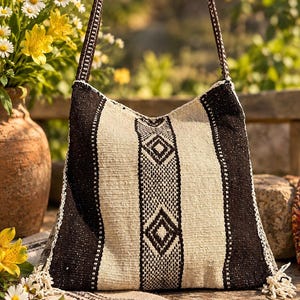 Könnte beinhalten: Eine gewebte Umhängetasche mit einem geometrischen Muster in Schwarz und Off-White, bestehend aus vertikalen Streifen und Rautenformen. Die Tasche hat einen langen, passenden gewebten Riemen und kleine dekorative Quasten an den unteren Ecken.