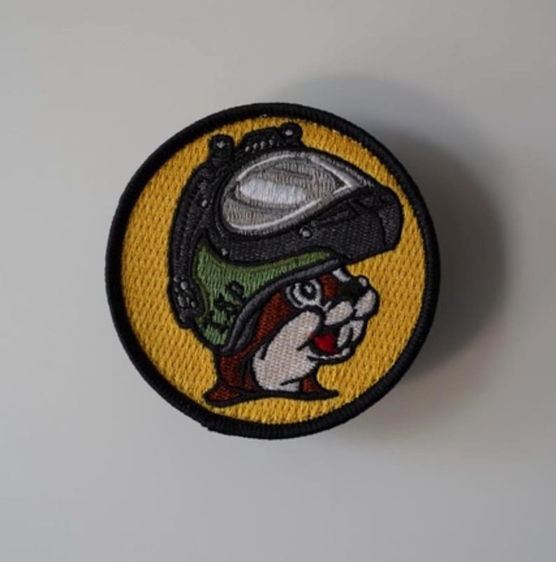 May include: Embroidered patch featuring a cartoon beaver wearing a pilot's helmet. The beaver has a red mouth and is set against a yellow background with a black border. The helmet is black, green, and silver.