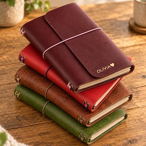 Peut inclure: Une pile de quatre carnets rectangulaires en cuir avec des fermetures élastiques. Le carnet supérieur est bordeaux foncé avec le nom OLIVIA et un symbole de cœur dorés. Les autres sont rouge, marron texturé et vert texturé, avec des œillets.
