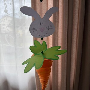 May include: A hanging Easter decoration featuring a white bunny head with a pink nose and a carrot. The carrot is orange with green leaves. The decoration is suspended by a thin string.