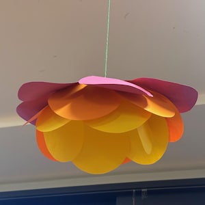 以下が含まれることがあります： ピンク、オレンジ、黄色系のグラデーションカラーの紙の花の吊り下げ飾り。緑色の紐で吊り下げられており、家の装飾に活気と遊び心を与えるアイテムです。