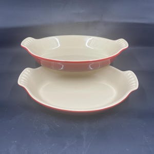 May include: Two oval-shaped ceramic baking dishes. The dishes have a cream-colored interior and a red rim. Each dish has two handles on either side. The dishes are stacked on top of each other.