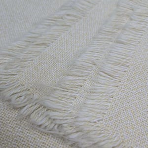 May include: Close-up of a cream-colored woven blanket with fringed edges. The blanket has a textured appearance and a subtle pattern. The fringe is soft and adds a decorative element to the blanket.